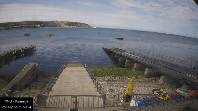 A view of a calm seaside scene with a small concrete pier extending into the water, surrounded by railings. To the right, there is a sloped ramp also with railings. On the rocky shore beneath the ramp, there are yellow lifeguard equipment and other items. Several small boats are anchored in the water, and a larger boat can be seen in the distance. In the background, there is a stretch of green land with cliffs along the coastline, and the sky is partly cloudy. The timestamp indicates the photo was taken on June 25, 2025, at 15:59.