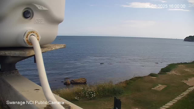 A view of the ocean with calm waves and a clear sky, taken from a webcam mounted on a ledge. Part of a white device with a cable connected to it is visible in the foreground on the left. Below the horizon, there is a grassy area with some small plants and a stone pathway. In the distance, a small boat is seen on the water, and a headland is on the right side. The timestamp on the top right reads "25-06-2025 16:00:00".