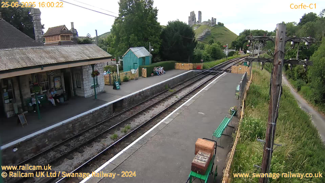 A small train station platform with a stone building on the left and a few people sitting on benches. There are some flower pots, a sign that says "Way Out," and a small teal-colored shed behind the seating area. The train tracks run diagonally across the image, and there are green benches along the platform. In the background, a hill with ruins on top can be seen, along with trees and a clear sky. A wooden utility pole with wires is on the right side of the platform.