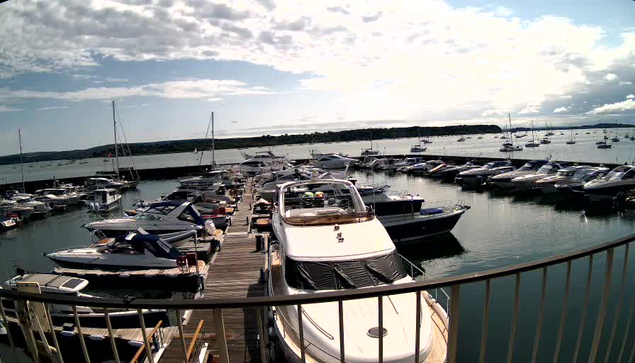 A marina filled with numerous boats docked along wooden piers. The boats vary in size and style, with some having sails and others being motorboats. The water is calm, reflecting the boats and the partly cloudy sky above. In the distance, there is a landmass with sparse vegetation. A metal railing runs along the foreground, suggesting the photo was taken from a balcony or walkway overlooking the marina.