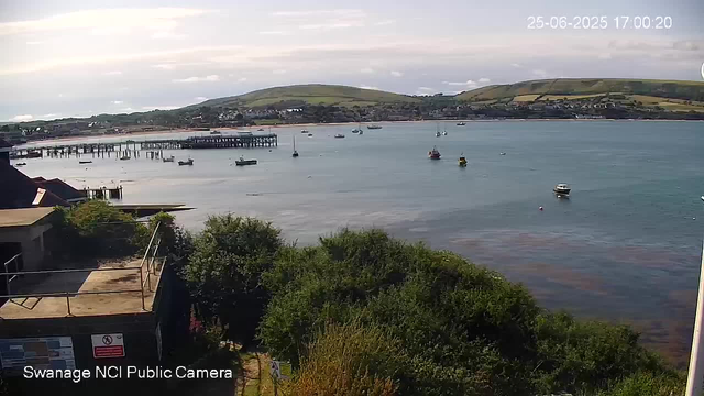 A coastal scene with calm waters and several small boats floating near the shore. In the background, there are green hills with patchwork fields and scattered buildings. The sky is partly cloudy with some sunlight breaking through. A wooden pier extends into the water on the left side of the image. The view is captured from a high vantage point, with trees and rooftops visible in the foreground. The timestamp in the upper right corner reads "25-06-2025 17:00:20".