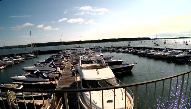 A marina filled with various boats and yachts docked along wooden piers, with additional boats floating on calm water. The sky above is partly cloudy with sunlight reflecting off the water's surface.