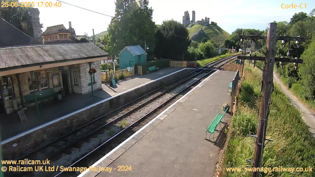 A small train station platform with a stone building on the left, featuring hanging flower baskets and a bench underneath a window. The platform is empty, and there's a sign indicating "Way Out." To the right, there are railway tracks with electric poles and wires, extending into the distance. Green benches are placed along the platform, and beyond the tracks, there are trees and a hill with the remains of a castle or ruins on top. The sky is clear, and the scene is brightly lit with sunlight.