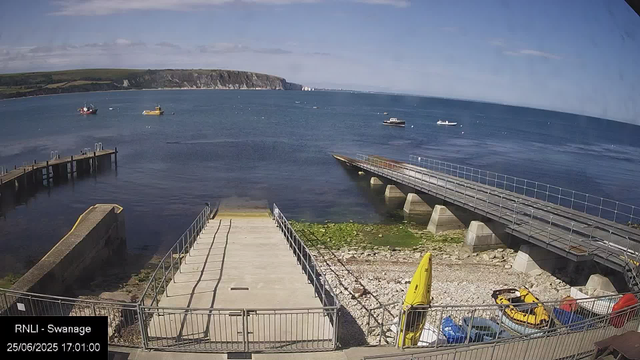 A view of a seaside dock area with several boats floating on the water. In the foreground, there are two docks extending into the water, with a small grassy and rocky area at the base. There are yellow rescue boats and other equipment on the ground near the fenced area. In the background, there are cliffs and a partly cloudy sky. The timestamp on the image reads 25/06/2025 17:01:00.