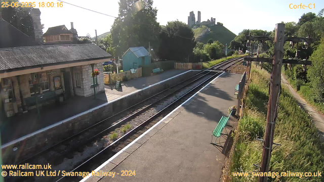 A small train station platform with a building on the left side featuring a corrugated roof and a hanging flower basket. Empty benches are positioned along the platform. A single railway track runs parallel to the platform, and there is greenery and trees to the right of the track. In the background, elevated ruins or structures are visible atop a hill, with a bright sky overhead.