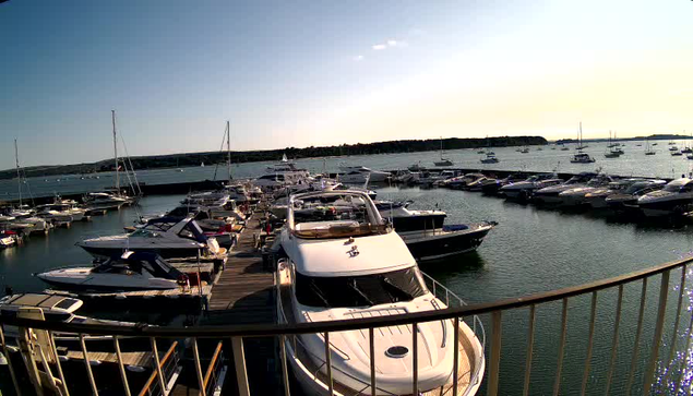 A marina filled with numerous boats and yachts docked along wooden piers. The water is calm, and the sky is clear with the sun setting or rising on the horizon. A curved railing is visible in the foreground.