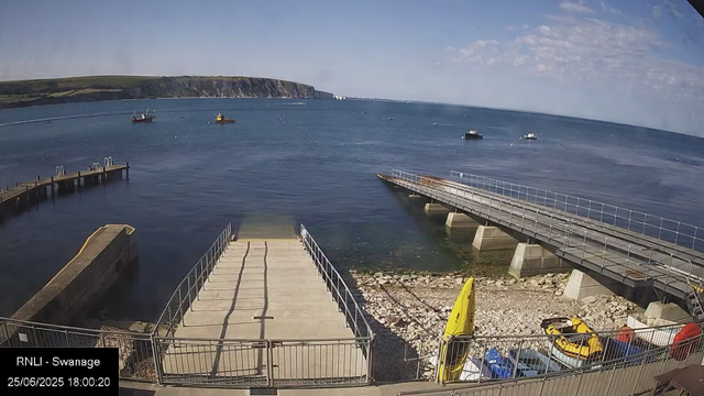 A view of a seaside harbor shows a concrete dock extending into calm blue water, with a small ramp leading down to the shoreline. Several boats are anchored in the distance across the water, and a landmass with cliffs and green hills is visible in the background. The sky is mostly clear with some scattered clouds. In the foreground, there are yellow safety floats and some parked cars below a metal railing. A timestamp in the lower left corner indicates the date and time as June 25, 2025, at 6:00 PM.