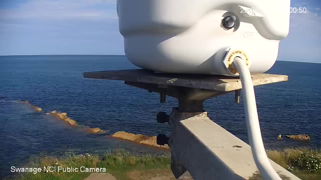 A webcam view overlooking a body of water with rocks along the shoreline. In the foreground, there is a white electronic device mounted on a metal bracket, connected by a white cable. The sky appears partly cloudy. The bottom left corner displays the text: "Swanage NCI Public Camera."
