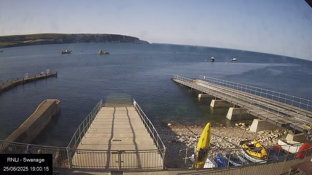 A view of a small harbor with a concrete pier extending into the water, surrounded by metal railings. Several boats are anchored in the distance on calm water. There are two yellow boats on the shore, along with some other smaller watercraft and equipment. The sky is clear with a distant landmass visible on the horizon. The timestamp indicates the date as June 25, 2025, at 19:00.
