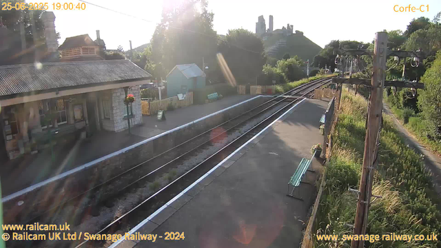 A view of a small railway station platform bathed in sunlight. There are two green benches, one visible near the bottom right corner and another further along the platform. A small building with a corrugated roof and flower baskets hanging outside is on the left. In the background, there are trees, a hill with ruins or old structures on top, and some buildings. To the right, there are railway tracks, and overhead electrical lines on wooden poles. The overall scene appears calm and quiet during late evening, with the sun shining brightly.