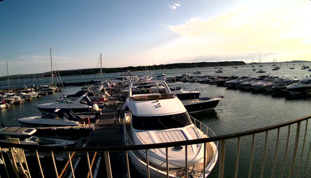 A marina with several boats docked along a wooden pier, extending into calm water. The boats vary in size, with the largest one in the foreground, and there are many more boats visible in the distance. The sky is partly cloudy with the sun setting or rising, casting a warm glow over the scene. A curved metal railing is visible at the bottom of the image.