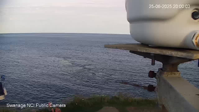 A view of the ocean with calm waves stretching out to the horizon under a slightly cloudy sky. In the foreground on the right, part of a large, gray cylindrical structure is visible, mounted on a rusted metal support. The top right corner shows a timestamp indicating the date as June 25, 2025, and the time as 20:00:03. The bottom left corner displays the text "Swanage NCI Public Camera."
