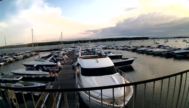 A marina filled with numerous sailboats and motorboats docked along a wooden pier, with calm water and a cloudy sky during sunset in the background.