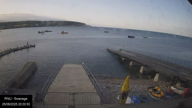 A view of a calm harbor with several boats floating on the water. A concrete pier extends into the bay, with a small yellow boat and some equipment on the shore. The background features distant cliffs and a cloudy sky. The timestamp shows the date as June 25, 2025, at 8:00:50 PM.