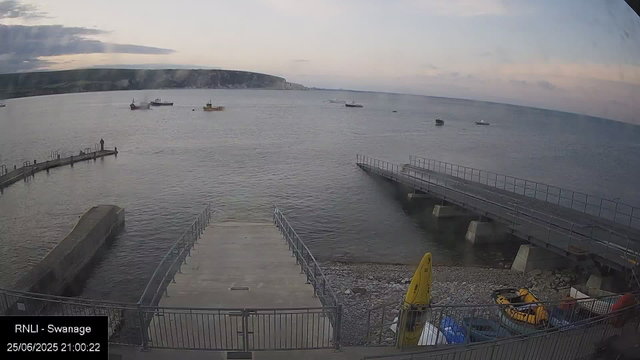 A view of a harbor with calm water and several boats anchored in the distance. There are two docks extending into the water, one of which appears partially submerged or damaged. In the foreground, there are yellow safety equipment and a few vehicles. The background shows a coastline with cliffs and a cloudy sky during dusk.