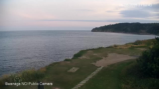 A scenic view of a coastal landscape at dusk: the calm sea stretches out to the horizon, with a gentle shoreline and a small grassy area with a stone pathway leading toward the water. In the background, a tree-covered headland extends into the sea. The sky is partly cloudy, with soft pink and blue hues. The image is timestamped "26-06-2025 21:00:43" and labeled "Swanage NCI Public Camera" in the lower left corner.