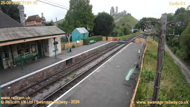 A small railway station platform with a stone building on the left, decorated with hanging flower baskets and green benches. There is a green shed behind the platform, surrounded by a wooden fence. Electrical wires run overhead, and the tracks extend into the distance towards a hill with castle ruins atop it. The station area is bordered by greenery and trees, with a narrow path visible on the right side. The overall scene is during daytime with overcast sky.