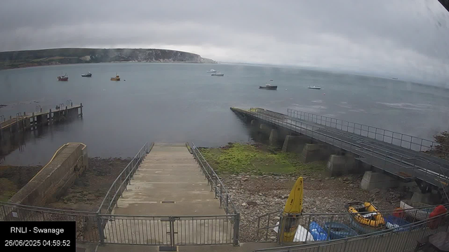 A view of a coastal area under grey, overcast skies. Several boats are anchored in calm water. There are concrete steps and walkways leading down to the water, with some green moss or algae growing near the shoreline. A yellow safety sign and some equipment are visible in the foreground, enclosed by a metal fence. The landscape in the background includes a distant cliff or hillside with patches of green vegetation. The timestamp indicates the image was taken early in the morning on June 26, 2025.