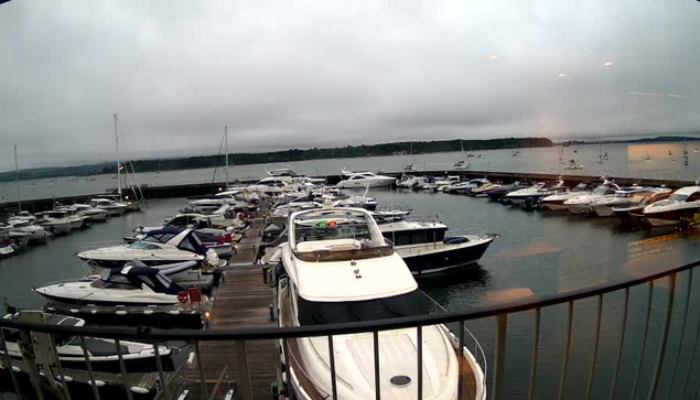 A view of a marina filled with various boats and yachts docked along wooden piers, with a calm body of water extending to the horizon under a cloudy sky.