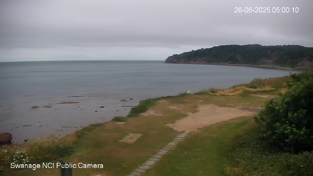 A view of a coastal landscape with a sandy and grassy shoreline leading to calm water, a tree-covered headland in the distance, and an overcast sky. The timestamp in the upper right corner reads "26-06-2025 05:00:10," and the bottom left corner indicates "Swanage NCI Public Camera."