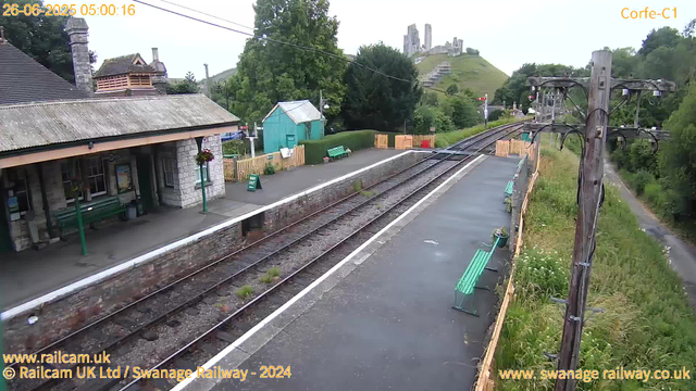 A small train station platform with a stone building on the left, featuring a porch with hanging flower baskets and green benches. Track lines run parallel along the platform, with a small green shed and a hedge in the background. Two green benches are placed on the platform, and there are several wooden fences and a sign that reads "WAY OUT". To the right, there's a tall wooden utility pole with electrical wires, and a narrow path runs alongside the platform. In the background, a hill with the remains of an ancient castle or ruins is visible, surrounded by lush green trees. The scene is captured early in the morning, as indicated by the timestamp.