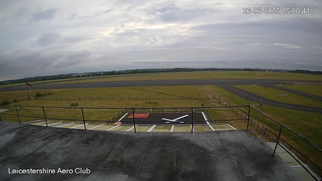 A view from a webcam at Leicestershire Aero Club showing an overcast sky with gray clouds. In the foreground, there is a rooftop with a railing, and a helicopter landing pad with a large white helicopter silhouette painted on it. The surrounding area includes grassy fields, a few small structures, and a runway in the distance. The timestamp in the upper right corner indicates the photo was taken on June 26, 2025, at 05:00:41.