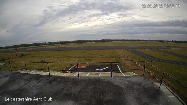 A view from a webcam at Leicestershire Aero Club shows an overcast sky with scattered clouds, a runway stretching across the horizon, and a tarmac area with a large helicopter landing pad in the foreground. The date and time displayed are 26-06-2025, 06:00:13.