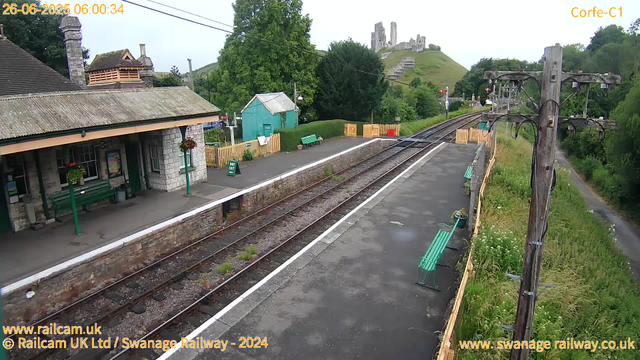 A small train station platform with a building on the left, featuring flower baskets hanging from the walls. There are green benches along the platform, a green shed in the background, and some colorful barriers at the far end. The railway tracks run through the center, with lush green trees and a grassy hill with ruins of an old castle on top in the background. A wooden utility pole with wires stands on the right side, and a narrow pathway runs parallel to the tracks on the far right. The scene appears to be early morning, as indicated by the timestamp.