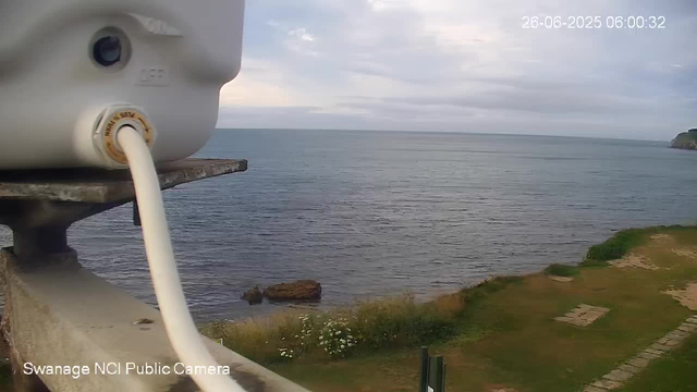 A close-up of a white camera mounted on a metal stand overlooking the ocean. The camera has a cable plugged into it, and the ocean stretches out under a cloudy sky with land visible on the right side in the distance. The timestamp in the upper right corner reads "26-06-2025 06:00:32," and in the bottom left corner, it says "Swanage NCI Public Camera."