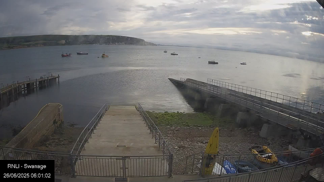 A view of the seaside at dawn with several boats anchored in the water. A stone pier extends into the sea, with a ramp leading down to the shore. The sky is cloudy, and the water has small ripples. On the shore, there is some green moss or algae, and various equipment and a yellow caution sign are visible near the bottom right corner. A timestamp indicates the image was taken on June 26, 2025, at 6:01 AM, and the location is labeled as RNLI - Swanage.