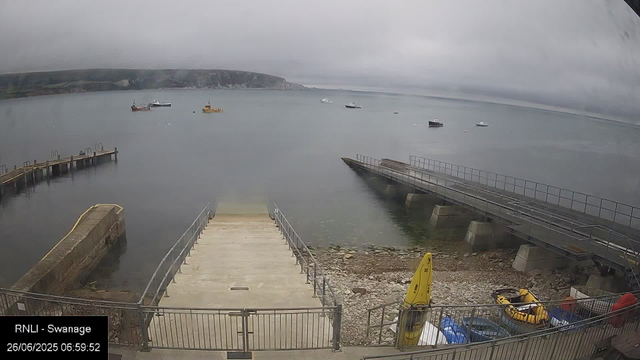 A view of a cloudy day at a harbor with several boats anchored in the water. There are two docks extending into the water, one on the left and one on the right. The shoreline has rocks and a small area with equipment and yellow safety gear. The scene appears calm with no people visible.