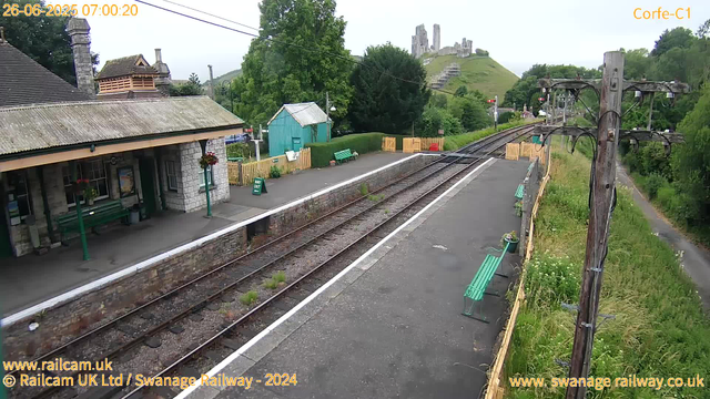 A small train station with a single platform and railway tracks running through it. There is a stone building with a sloped roof on the left, decorated with hanging flower baskets. Two green benches are situated along the platform. In the background, there is a green shed, a yellow fence, and lush trees surrounding the area. A castle or fortress is visible on a hill in the distance. To the right, there is a wooden utility pole with wires, and a narrow path runs alongside the platform through tall grass and wildflowers. The image has a timestamp of June 26, 2025, at 7:00 AM.