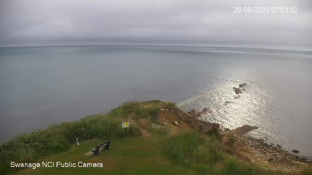 A view of a coastal scene on a cloudy day, showing a rocky shoreline extending into the ocean. The ocean stretches into the distance under a gray sky, with sunlight reflecting off the water. There is some greenery and a bench at the edge of a small grassy area in the foreground. The image is timestamped June 26, 2025, at 7:01 AM, and has a label indicating it is from the Swannage NCI Public Camera.