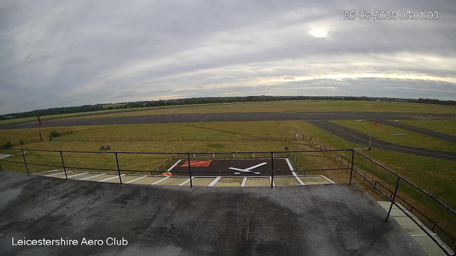 A view from a webcam at Leicesterhire Aero Club showing an overcast sky with some sunlight breaking through clouds. The image includes a runway in the distance, a grassy area with a bench and a small flag pole, and a helipad in the foreground with a white helicopter tail rotor blade lying on the ground. The image also has a timestamp in the upper right corner showing "26-06-2025 07:01:03," and the bottom left corner displays the text "Leicestershire Aero Club."