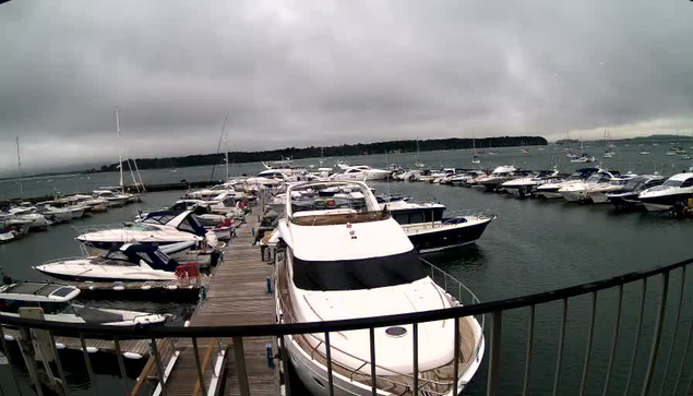 A marina filled with numerous boats and yachts docked along a wooden pier. The water is calm, and the sky is overcast with dark clouds. In the background, there is a distant shoreline with trees. A black railing is visible in the foreground, indicating that the photo was taken from a balcony or similar vantage point.
