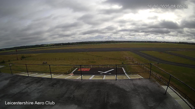 This is a wide view of an airfield on a cloudy day, with the tarmac and a small helipad marked with a white "X" in the foreground. There is a wooden railing around the helipad area, and a small red and black painted section with a white cross nearby. The runway stretches across the middle distance, with grassy areas on either side, and a distant horizon with trees and low hills under a cloudy sky. The timestamp shows it was taken on June 26, 2025, at 8:00 AM. The text "Leicestershire Aero Club" appears at the bottom left corner.