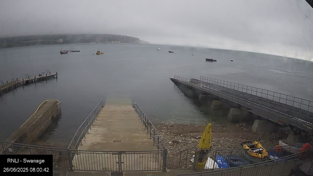 A view of a harbor on a cloudy day, with several boats floating on calm water. There is a concrete pier extending into the water in the foreground, and a ramp or slipway leading into the sea. On the right side, some equipment and a yellow lifebuoy are visible, along with a gravel area. The background shows distant cliffs partially obscured by fog or low clouds. The timestamp indicates the image was taken early in the morning on June 26, 2025.