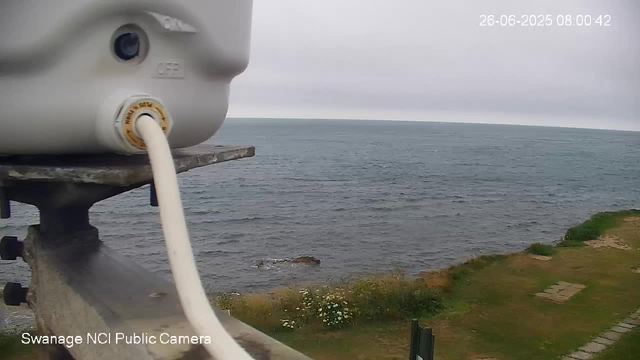 A webcam captures a view of the ocean on a cloudy day, with the horizon in the distance. In the foreground, part of the camera housing and a cable are visible, mounted on a weathered metal railing beside a grassy area with some small white flowers. The timestamp on the image reads "26-06-2025 08:00:42," and the label indicates it is from a "Swanage NCI Public Camera."