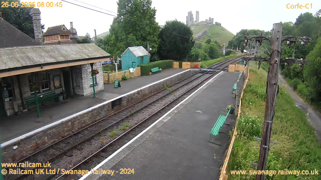 A small train station with a single platform featuring benches, a green shed, and a surrounding lush green landscape. There are overhead power lines and railway tracks running through the station, with a hill in the background that has a castle ruin on top. The scene appears to be early in the morning on June 26, 2025, as indicated by the timestamp. The station is quiet with no visible passengers.