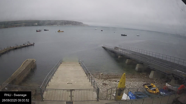 A view of a harbor on a cloudy day with several boats anchored in the water. In the foreground, there are two piers extending into the water, with one being a small concrete slipway and the other a metal walkway. A yellow lifeguard board is positioned near the edge of the small slipway, and some equipment and containers are stored behind a safety fence. The background shows a distant coastline with cliffs under a gray, overcast sky.