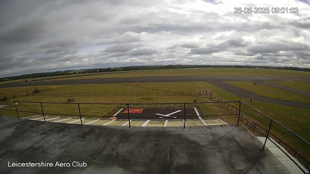 A view from a webcam at Leicesterhire Aero Club showing an overcast sky with thick clouds. The foreground features a small black helipad with a white letter "H" and an orange outline, surrounded by a metal railing. There is an empty parking area below with a handicap parking space visible. In the background, there are grassy fields, a runway, and a distant tree line under the cloudy sky. The timestamp on the image reads "26-06-2025 09:01:03."