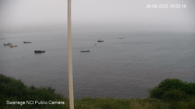 A foggy day over a body of water with several anchored boats scattered across the surface. There is some greenery in the foreground and a white vertical pole near the center of the image. The timestamp in the upper right corner reads "26-06-2025 10:00:18," and the bottom left corner indicates "Swanage NCI Public Camera."