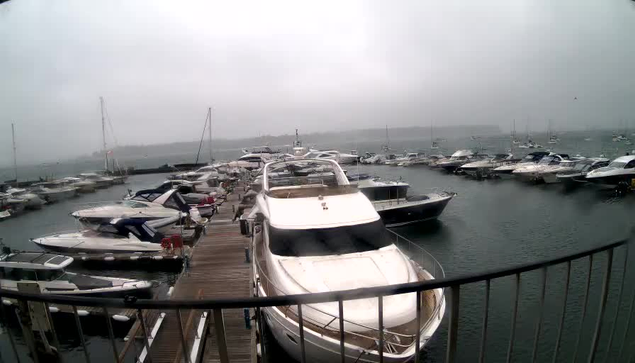 A marina filled with numerous yachts and boats docked along wooden piers under an overcast sky, with sails visible on some boats and calm water reflecting the vessels.