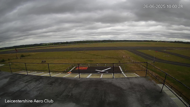 Overcast sky with dark gray clouds, view of an airfield with runways stretching into the distance and a helicopter landing pad with a red and yellow marking in the foreground. The image is from a webcam at Leicestershire Aero Club, timestamped 26-06-2025 at 10:00:28. The horizon shows open fields and distant trees.