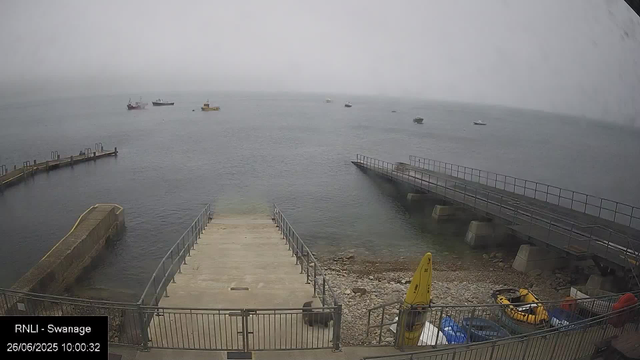 A view of a harbor with a concrete pier extending into calm waters. Several boats and ships are anchored in the distance on a foggy day. There is a small yellow buoy and some equipment, including a yellow kayak and a blue water container, near the shore. The overall atmosphere appears misty and overcast.