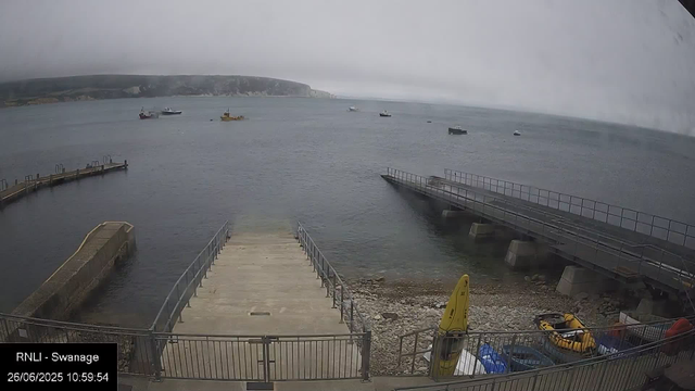 A view of a harbor on a cloudy day, showing multiple boats floating on the water. In the foreground, there are two piers extending into the water, with one having a set of stairs leading into the sea. There's a small concrete platform at the water's edge, along with a shoreline of rocks and pebbles. In the lower part of the image, there are yellow safety signs, a yellow inflatable boat, and various other equipment. The background features a distant landmass with cliffs and some fog or low clouds obscuring the sky. The bottom left corner shows the text: "RNLI - Swanage 26/06/2025 10:59:54."