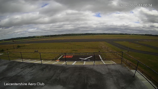A view of the runway and surrounding airfield at Leicestershire Aero Club under a cloudy sky, with some patches of blue, and a windsock on the left side. The photo is timestamped June 26, 2025, at 11:00 AM. In the foreground, there is a small helipad with a white helicopter silhouette painted on it. The scene is empty with no aircraft visible on the tarmac or runway.