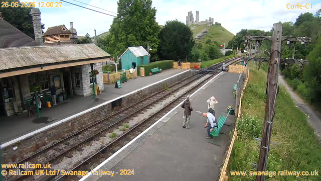 A small train station platform with a building on the left, several people waiting and chatting, some sitting on green benches. There are yellow fences at the end of the platform and a small teal-colored shed. Overhead, electrical wires run above the tracks. In the background, there are lush green trees, a path running parallel to the tracks on the right, and a hill with the remains of an old castle or fortification on top of it. The image was taken at midday, and the sky is partly cloudy.
