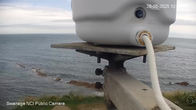 A webcam captures a view of the ocean with a cloudy sky overhead. In the foreground, there is a close-up of a white metal device mounted on a wooden platform supported by a rusty metal bracket. A white cable is connected to the device, and part of the platform is visible. The camera's date and time display in the upper right corner shows "26-06-2025 12:". The bottom left corner indicates that the source of the footage is from "Swanage NCI Public Camera."