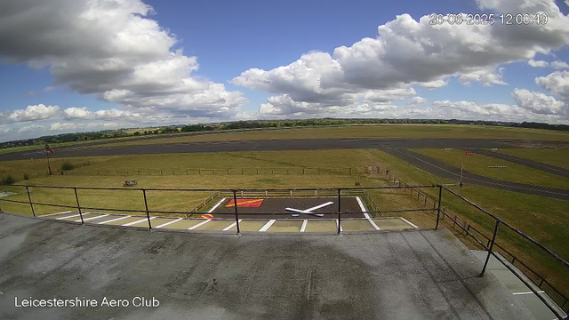 A view from a webcam at Leicester Aero Club showing an empty aircraft parking area with designated spots marked by white lines. Beyond, there is a grassy field and a runway, with a partly cloudy sky overhead. The timestamp in the top right corner indicates the date and time as June 26, 2025, at 12:00:49.